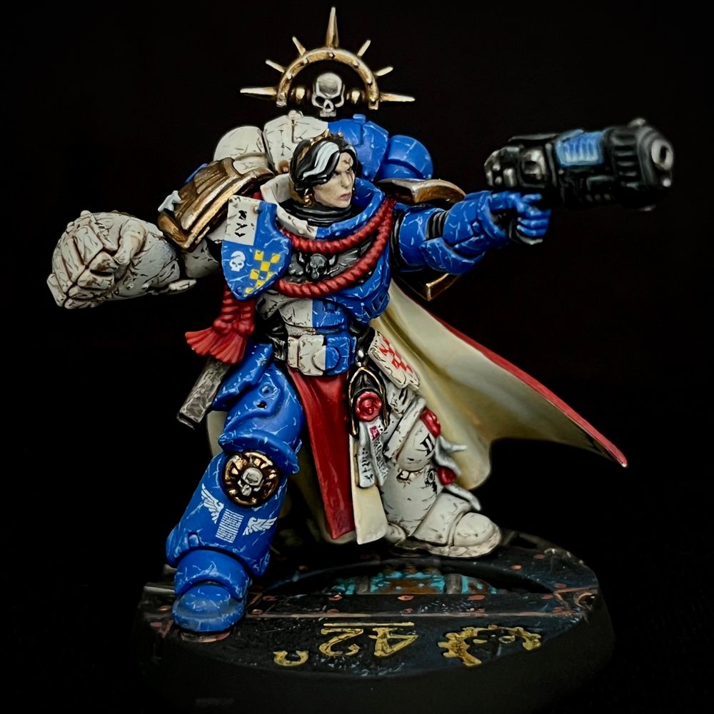A Novamarine Space Marine Captain equipped with a Power Fist and Plasma Pistol. She is wearing quartered bone and blue power armour, a red and cream cloak and is adorned with a lot of decoration and heraldry. She has black hair with a white stripe falling over one eye. All her equipment is battered and worn.