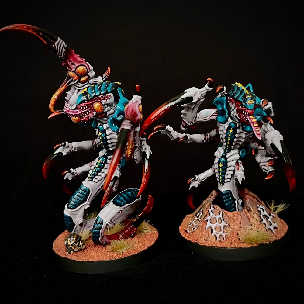 A pair of Hyper-Adapted Raveners painted with glossy black/blue carapace with yellow markings, white/purple skin with red markings, and dark red claws and spikes. One has bright orange poison-filled sacs on its face and claws, while the other has a number of additional yellow eyes on its head and down it’s tongue
