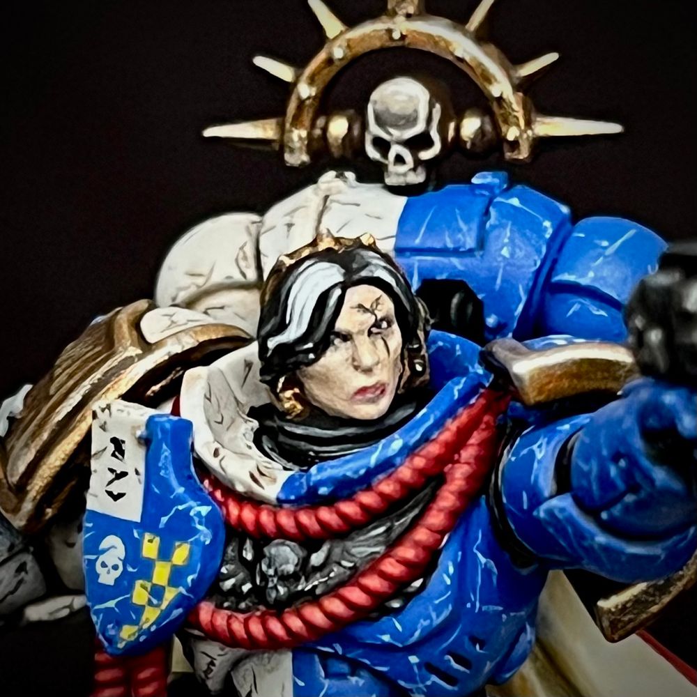 A Novamarine Space Marine Captain equipped with a Power Fist and Plasma Pistol. She is wearing quartered bone and blue power armour, a red and cream cloak and is adorned with a lot of decoration and heraldry. She has black hair with a white stripe falling over one eye. All her equipment is battered and worn.