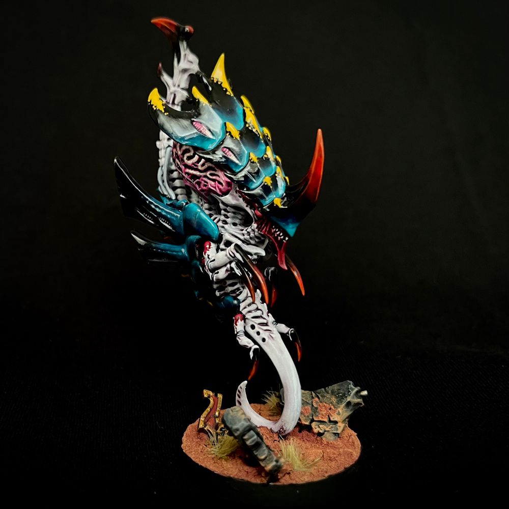 A Tyranid Neurothrope with black and turquoise carapaces with yellow markings, whitish purple skin with red markings, and oversized glowing pink brains. It has a large red horn at the front of its head