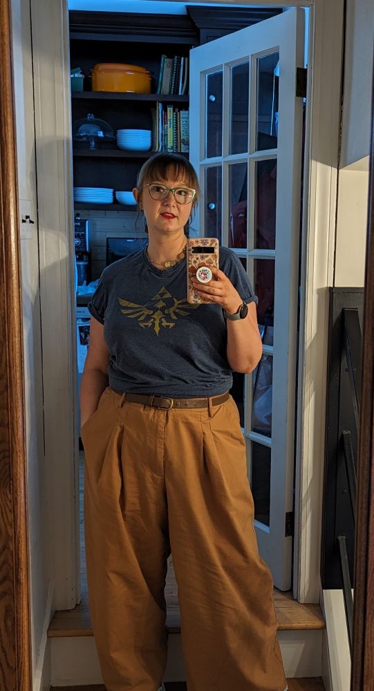 Mirror selfie of brunette woman with bangs, a ponytail, green and gold cat eye glasses, red lipstick, and a blue t-shirt with a yellow Zelda logo across the front, and khaki coloured wide-leg pants