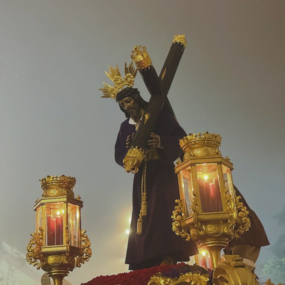 Photograph of a life-sized, three dimensional Christ figure carrying a cross, silhouetted against a misty grey dawn sky. The photo is from a Spanish Easter procession. In the foreground there are two ornate gold lantern-style lamps, with candles inside, which are part of the float that the statue is atop.  