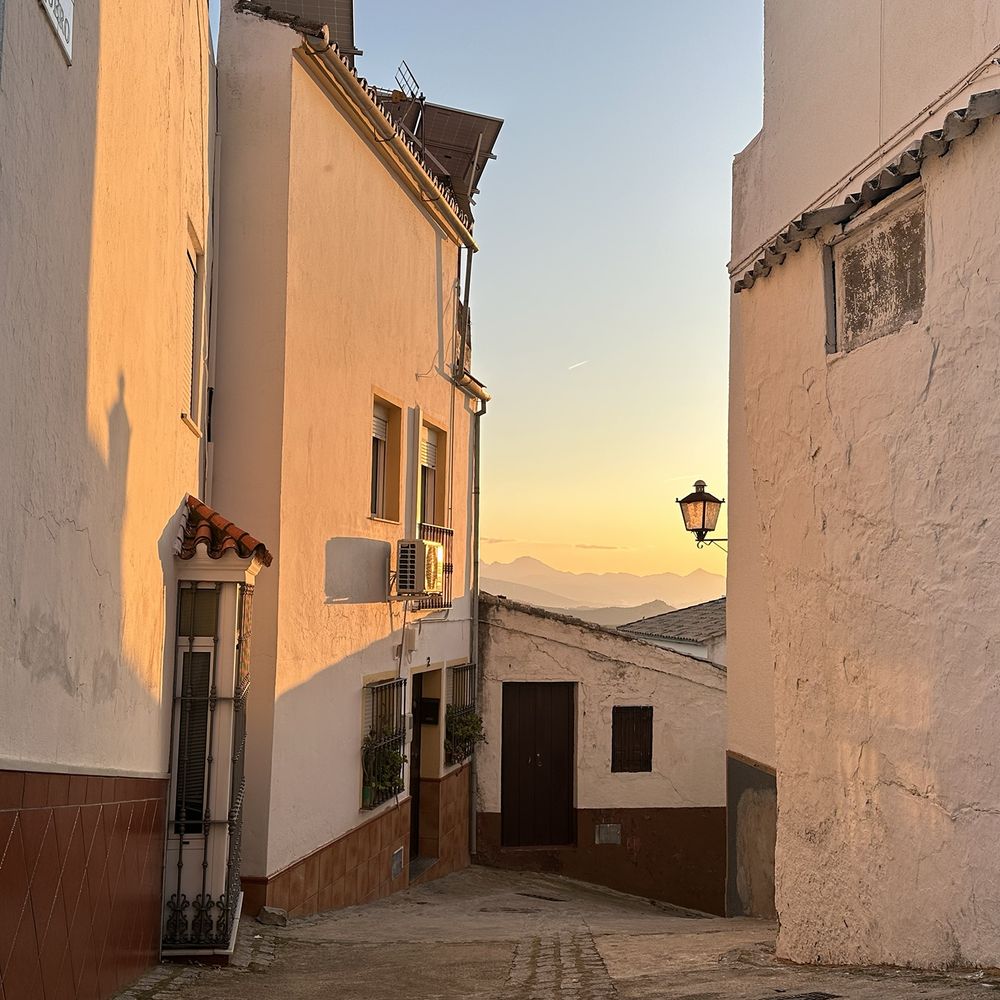 Square photo of a quiet Spanish village corner at sunset. The light is rosy orange on the square white house fronts. In the distance there are mountains.