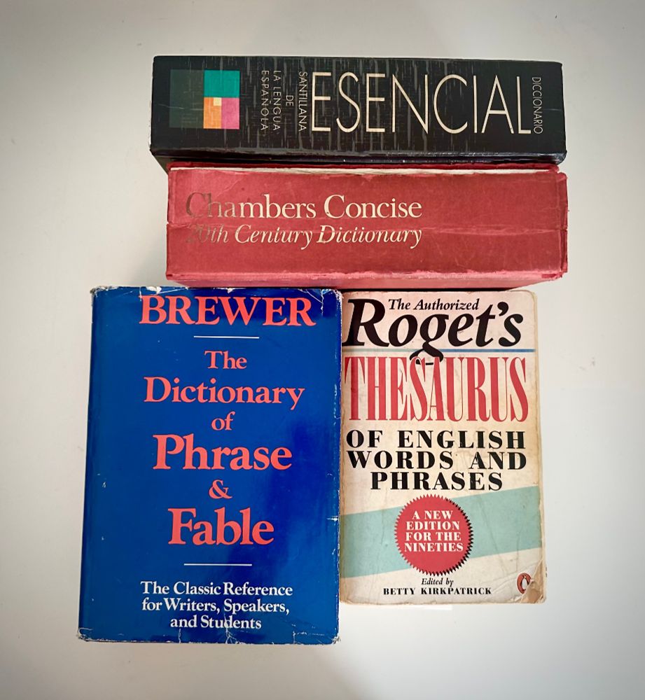 Square photograph of four shabby books. The front covers of two are shown, above them are the horizontal spines of two more. All of the books look well used.
At the very top is a book with a black spine and a small coloured geometric publishing house symbol. The title of the book is in white sans serif type and reads “Diccionario Esencial Santillana de la lengua española”. The word “Esencial” is in very large font, all caps. It is printed from left to right in the image (therefore from bottom to top of the book spine in reality.) It takes up about half the length of the spine. The other words are all in small caps and written horizontally across the spine.
The other horizontal book spine is a dull orange-red. The title is shown in a serif font in gold as “Chambers Concise 20th Century Dictionary”. 
The book on the left has a dark blue dustcover with big red type showing author and title “BREWER ı The Dictionary of Phrase & Fable”. The subtitle is in smaller white type: “The Classic Reference for Writers, Speakers and Students”.
To its right is a slightly smaller book with a mostly white cover with a mix of black and red type in a mix of fonts. The cover text reads: “The Authorized Roget’s Thesaurus of English words and phrases”. Then there is a red medallion-style ornament with the text, “A new edition for the nineties” in capitals, and below that the text reads “edited by Betty Kirkpatrick”. The logo of Penguin publishing house is visible in the very bottom right of the cover, but is partially obscured as the book is well-thumbed and the bottom right-hand corner is ragged.
