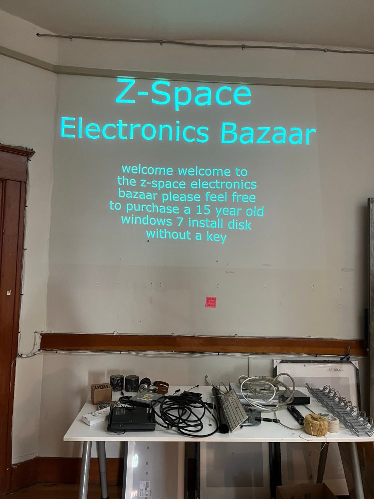Landing area on third floor. There is a table with stuff on it. 

A projector projects gently waving text on the wall

“welcome welcome to the z-space electronics bazaar please feel free to purchase a 15 year old windows 7 install disk without a key”
