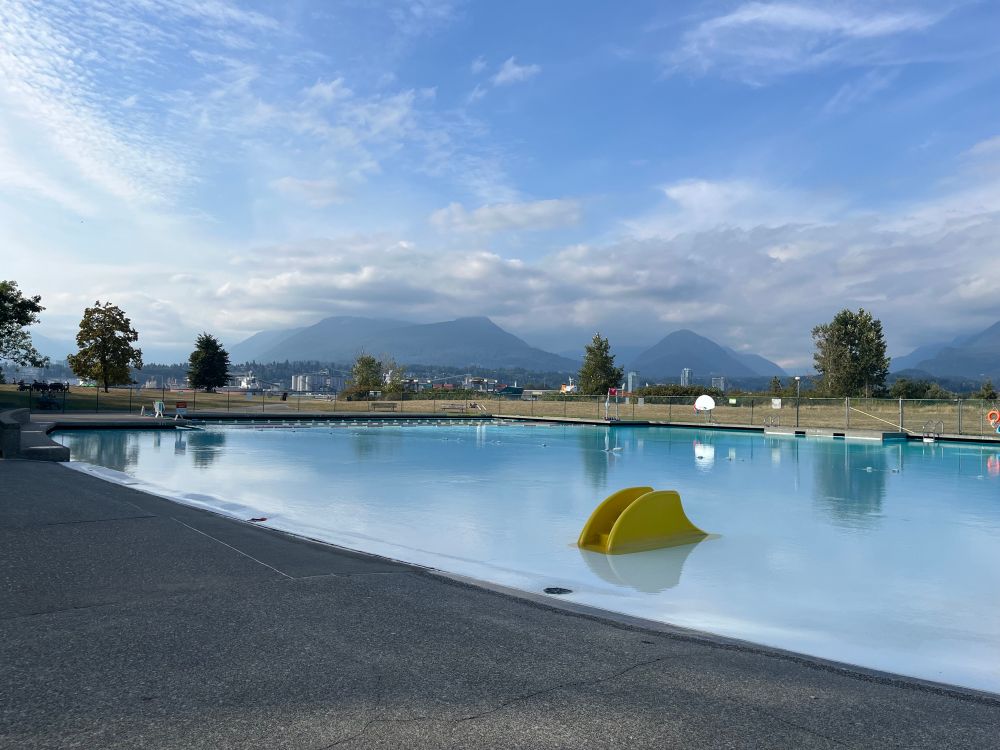 Evening across New Brighton Pool, no one in it. North Shore mountains in the distance. High clouds and sun. 