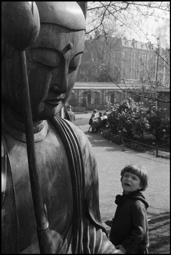 A young child gazes up at a tall statue of Buddha in a town center. Trees, apartment buildings, and shop fronts are in the distance. Photograph is attributed to Leonard Freed, with no date or location
