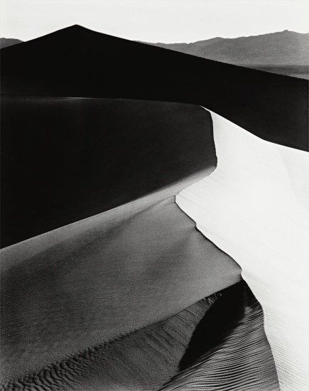 Curves of sand dunes are layered into the distance. As the sun rises over the top of one, the light glows in contrast to the remainer in shadow