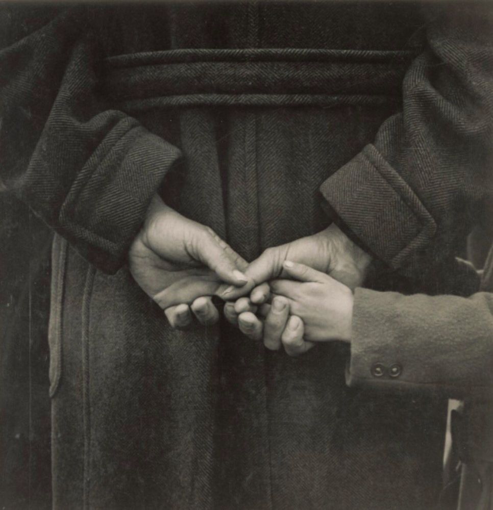 back of an individual in a long wool coat showing their hands clasped together and holding the smaller hand of a child standing close