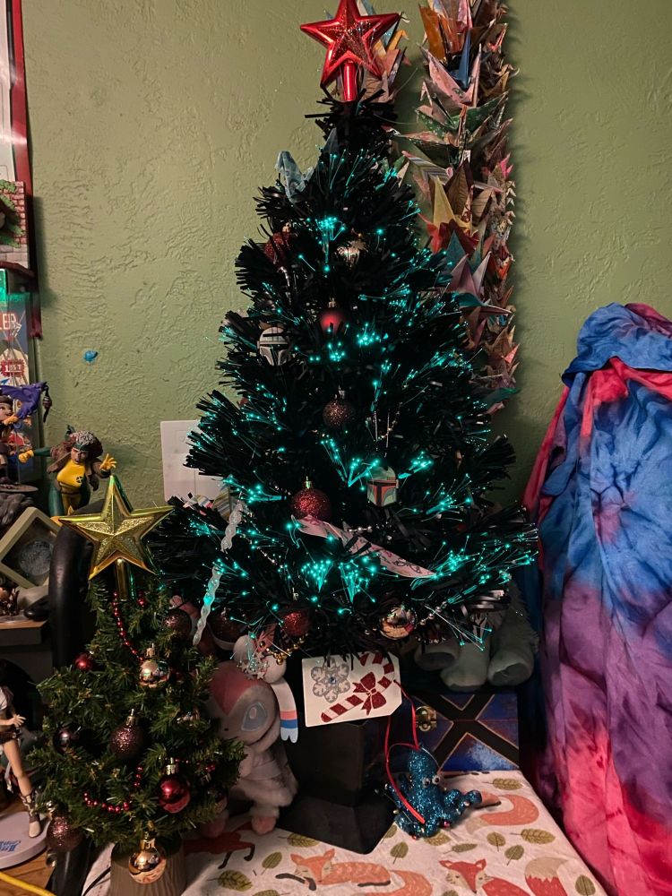 A small, black tabletop Christmas tree with fiber-optic lights, currently glowing teal. There are ornaments and origami decorating it.
There is an even tinier green tree beside it, also decorated with ornaments.