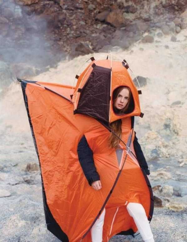 A woman stands, body covered by an orange tent, arms and legs poking through slits. Her head is covered by a smaller orange tent, face showing through a cutout.