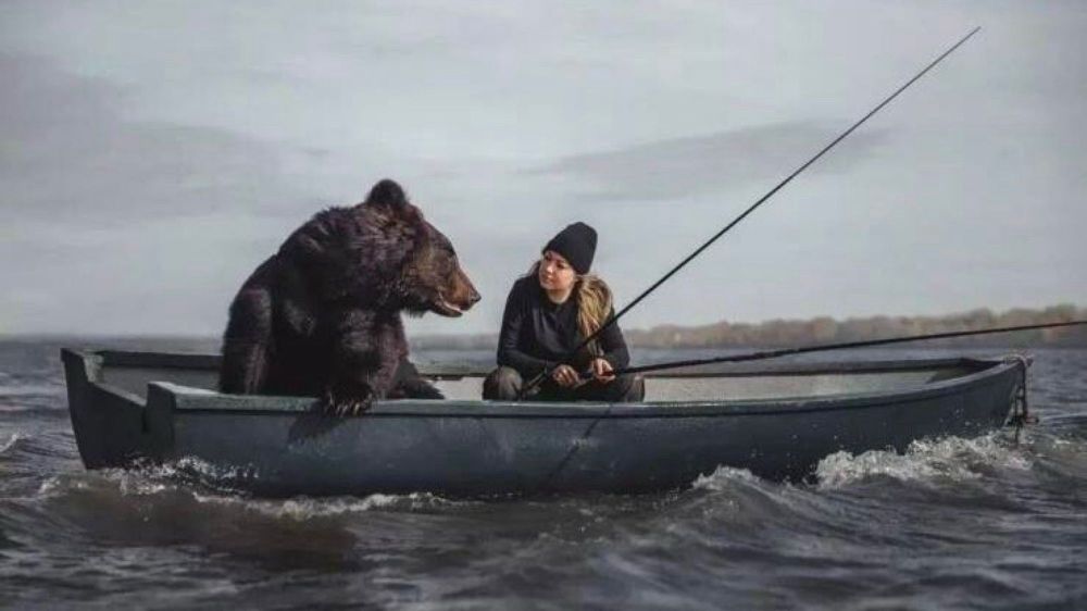 A woman and a bear sit in a boat, fishing.