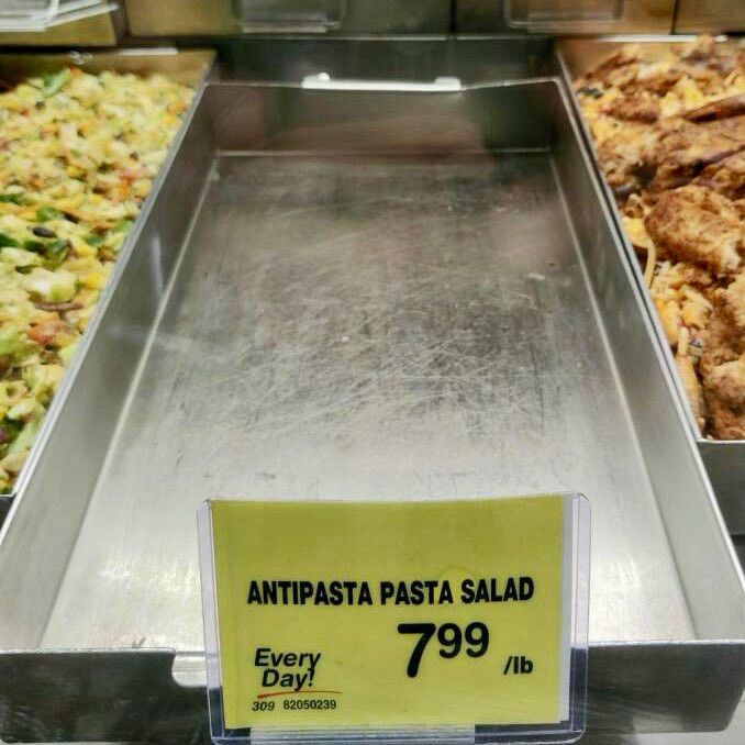 Empty food tray with a label reading "Antipasta Pasta Salad, $7.99 /lb" next to trays with food at a self-service counter.