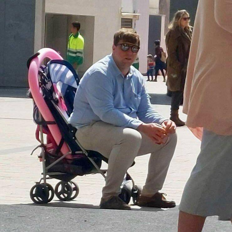 A man seated on a pink stroller, outdoors on a sunny day. Other people are visible in the background.
