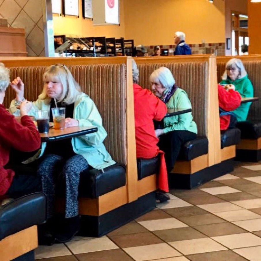 In three consecutive booths, three visually-similar women in turquoise tops sit across from three visually-similar women dressed in red.
