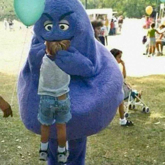 In a park setting, someone in a Grimace costume lifts a small child. The child’s head is in Grimace’s mouth.