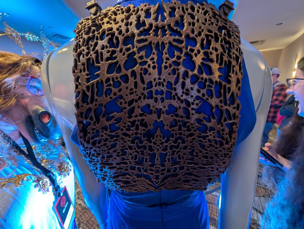 A closeup of the back of Moiraine's armor, intricately carved leather made to look like metal or other durable materials. To the left, is a person (ess) wearing a horse mask because they're bela the creator lol