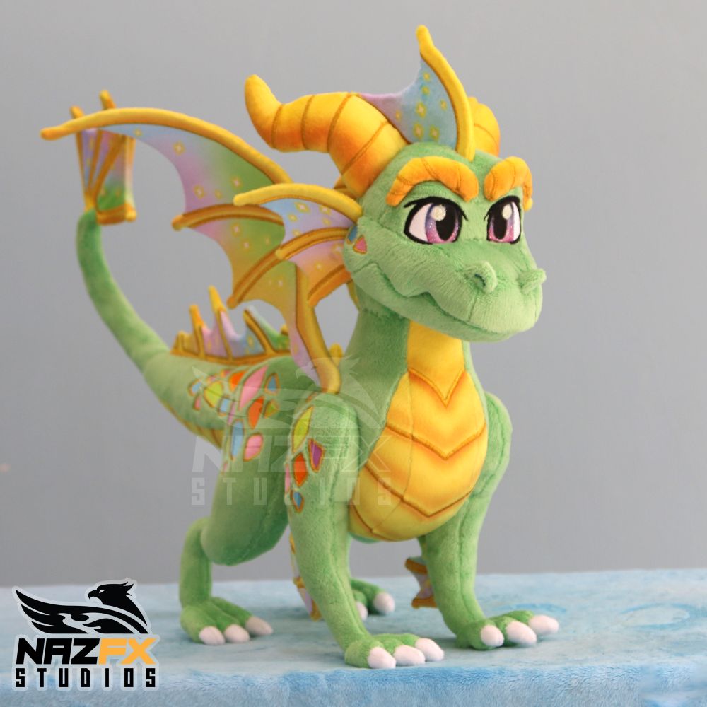 A handmade dragon plush. It is green with gold accents. It has frills with gold and pastel rainbow gradients. The eyes sparkle with glitter. Scales grace its sides in a rainbow of color. 