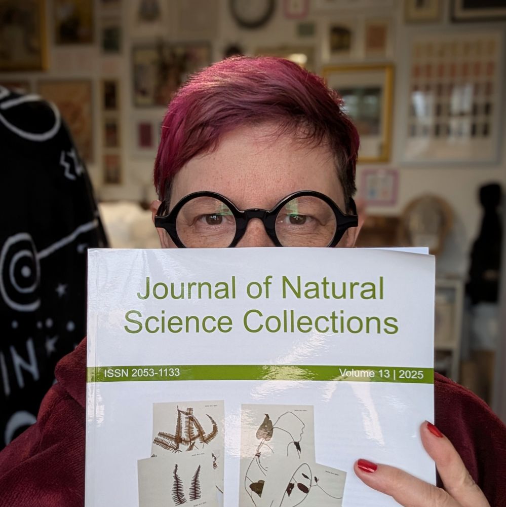 A middle-aged woman with short pink hair and round black glasses is peeping over a printed journal - it's Issue 13 of the Journal of Natural Science Collections