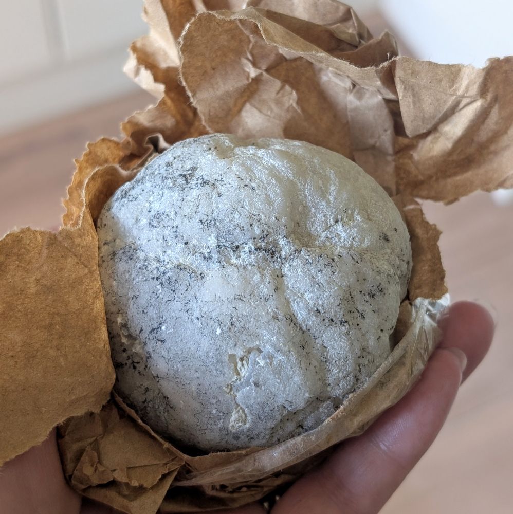 A close up view of a tennis ball sized round piece of rock. It is pale grey, and the surface is slightly bumpy. It's sitting on a layer of brown paper, which is torn to reveal the rock. It is being held up in the palm of a hand. 