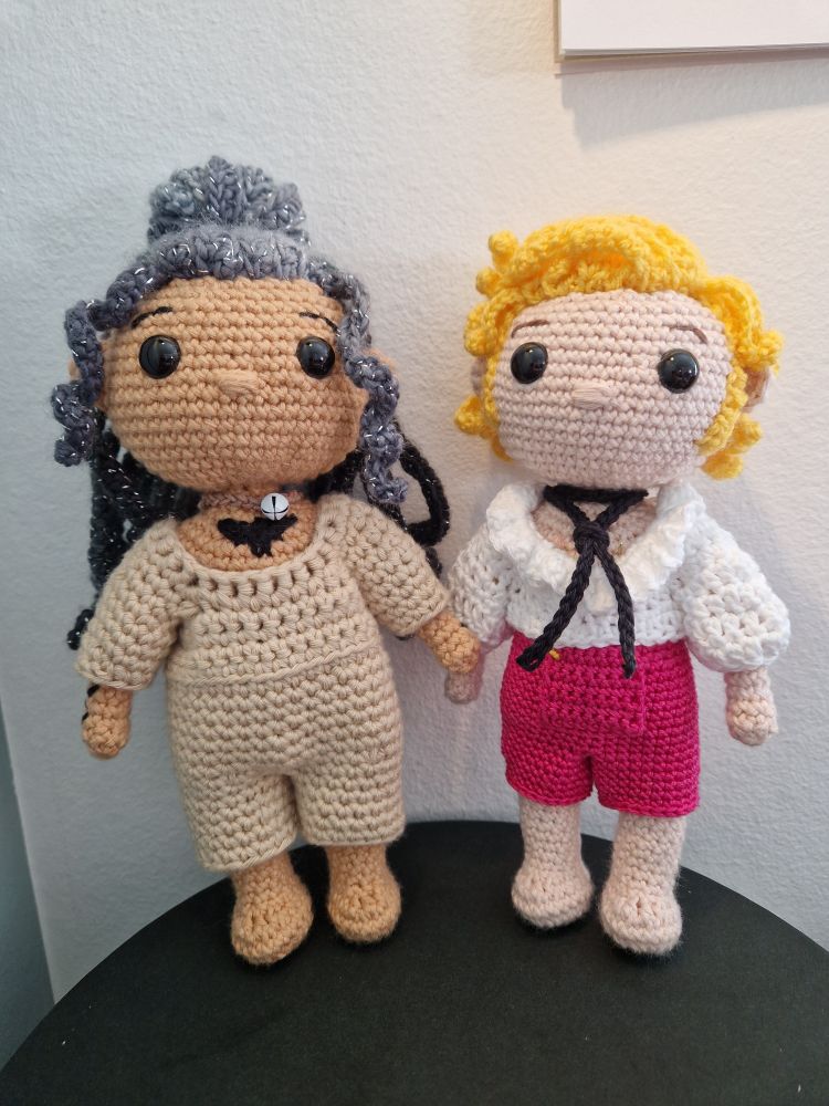 Two crocheted dolls/amigurumi of Ed and Stede from OFMD. Ed is in a beige onesie and cat collar and Stede is in pink breeches, a ruffled white shirt, and black cravat.