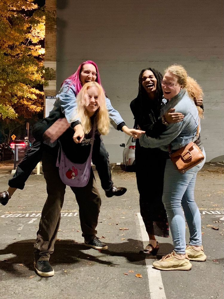 4 adults on the left, a person with pink hair on the back of a woman with blonde hair 

To the right Tuesday and her wife are embracing each other and smiling widely 