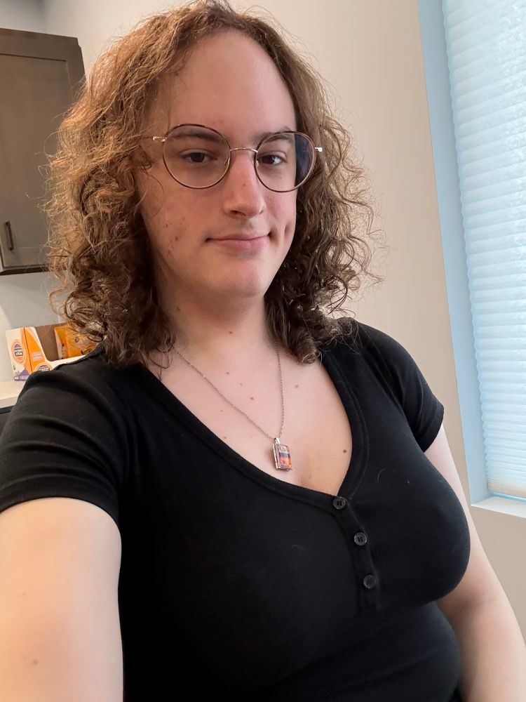 Selfie where I am wearing a black, low-cut top, my Dune book cover locket necklace, and my hair pretty curly from having washed it just a couple days ago. My glasses are new: round and large-ish, with thin brown rims. 