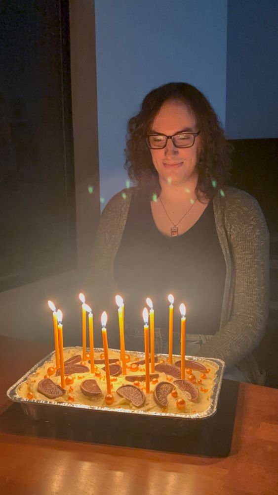 Me in a dark room, sitting at a table with lit candles in a birthday cake in front of me. I am very cute and wearing a moderately low-cut, black top with a thin, grey cardigan over my shoulders. I have my hands in my lap and I look extremely cute as I excitededly make a wish before blowing the candles out. 