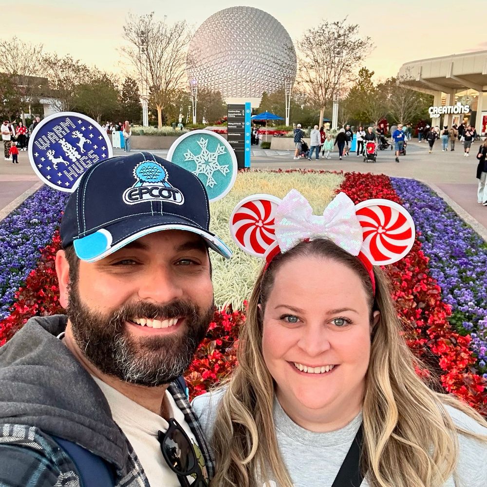 Chris and Kristy are smiling in front of a flower bed with Spaceship Earth in the background. He is wearing a blue EPCOT hat with white and blue, winter-themed mouse ears that’s says Warm Hugs and she is wearing red-and-white peppermint candy themed mouse ears and a sparkly white bow. 