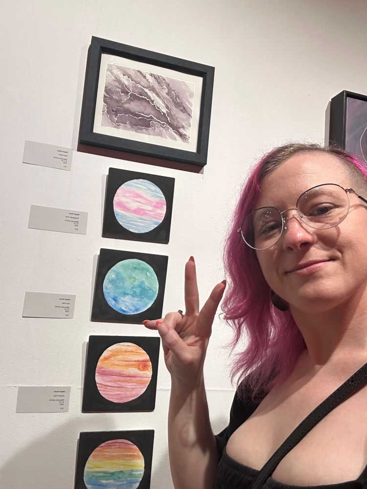 A white person with pink hair and glasses wearing a low-cut dress makes the peace sign while standing next to a series of vertically arranged watercolor paintings 