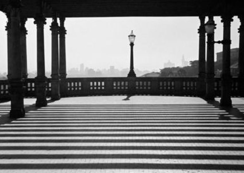 Foto em preto e branco de um pergolado com sombra e luz deixando marcas listradas no piso, um parapeito no fundo com um único poste de iluminação do estilo antigo a gás com o skyline da cidade de prédios ao fundo