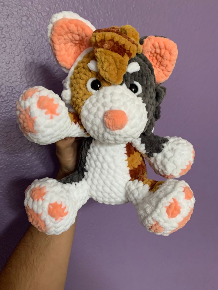 A calico cat plushie with a pink nose, paw pads, and inner ears. There is hair-like tufts of crochet atop the head