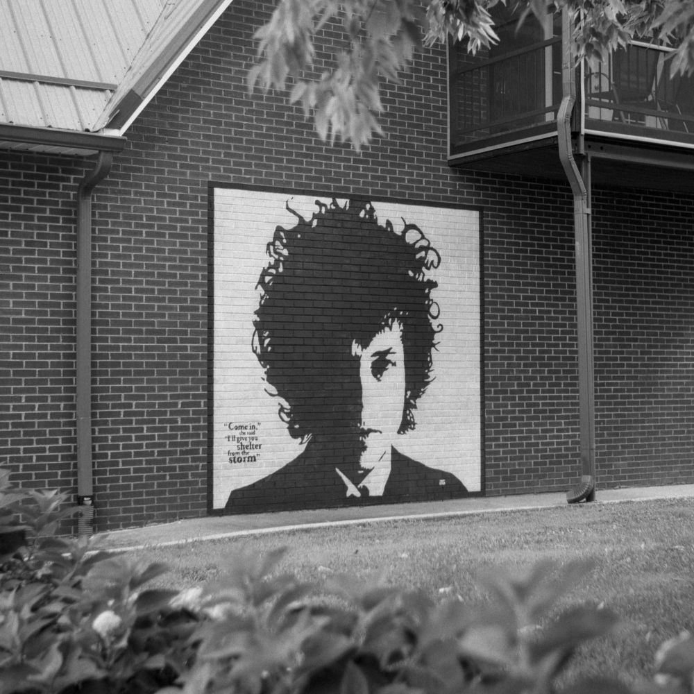 A black and white film photo. A large painted image of a young Bob Dylan is seen on a brick wall in Berea, Kentucky. Friday, July 18, 2025.