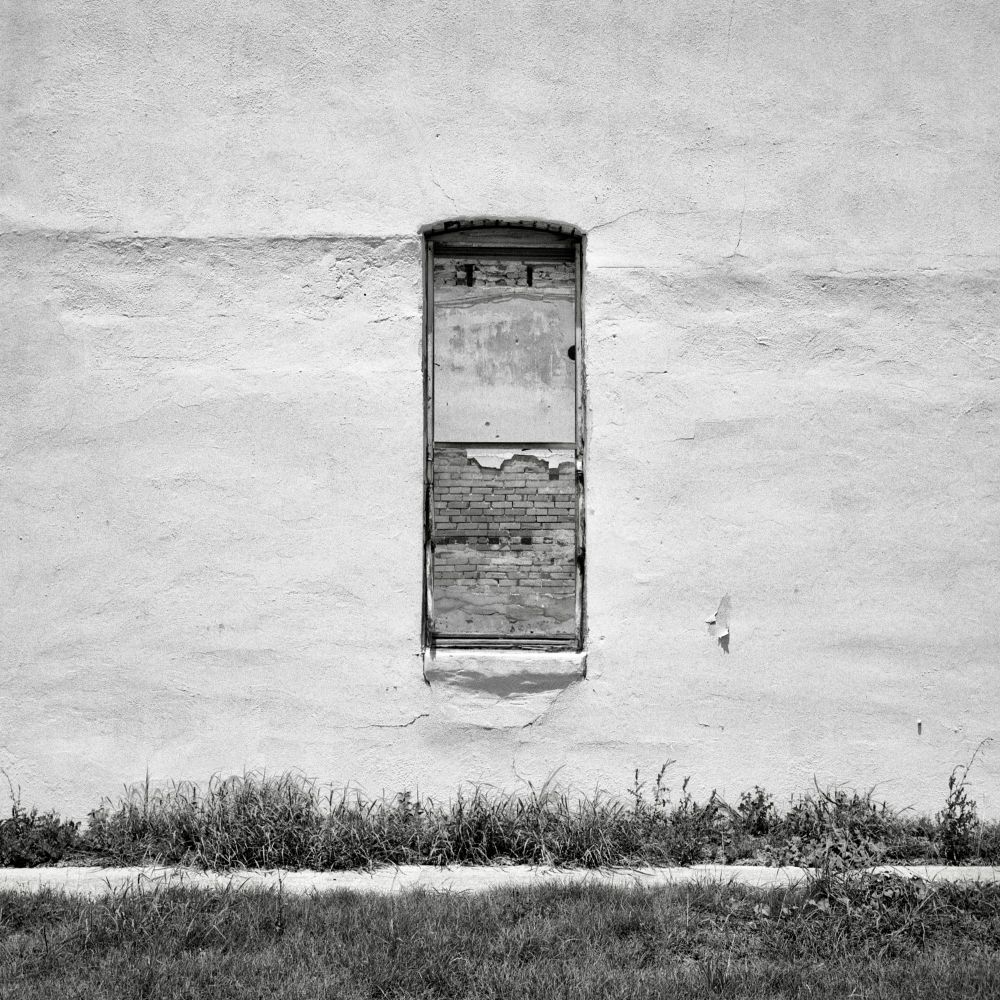 A black and white film photo. A narrow window on the side of an old abandoned building reveals the interior brick wall in Bartlett, Texas. Sunday, June 15, 2025. 