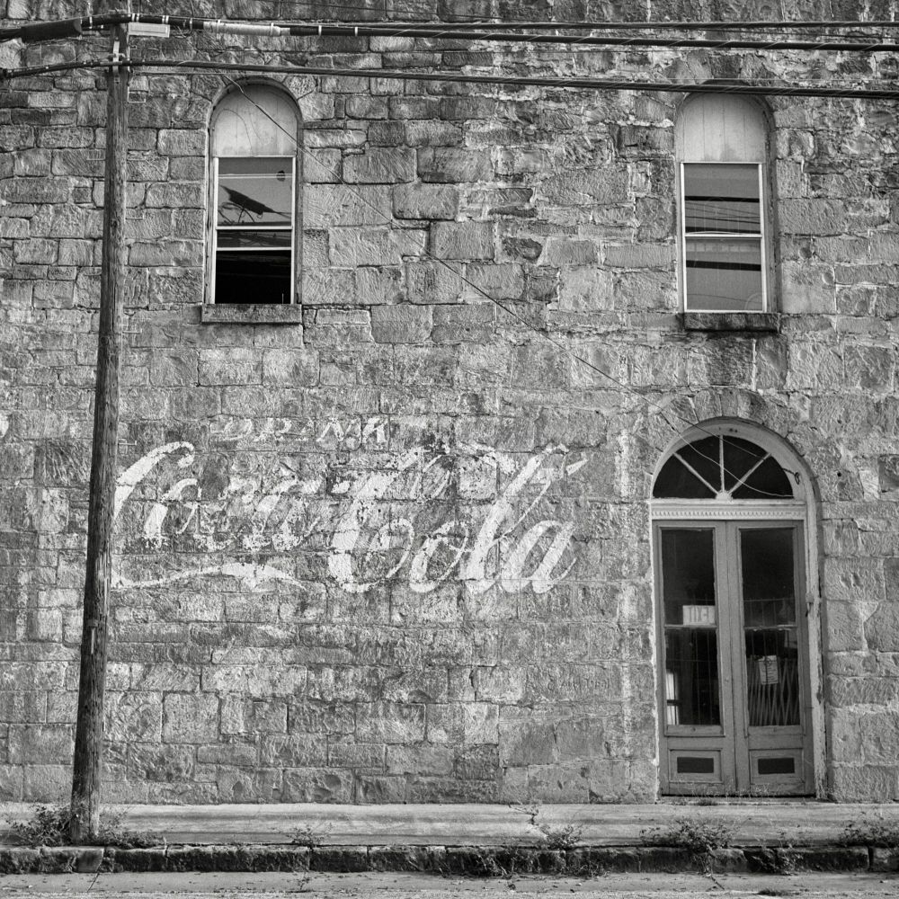 A black and white film photo. A weather-worn hand-painted sign that states “Drink Coca-Cola“ is seen on a very old stone building façade in Gonzales, Texas. Sunday, June 1, 2025.