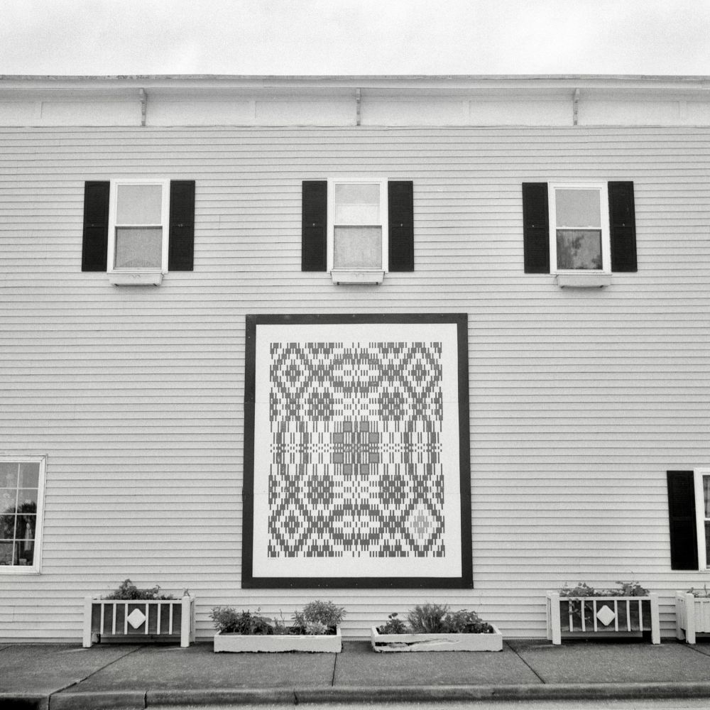 A black and white film photo. A large depiction of a quilt pattern is affixed to the side of an old house in Berea, Kentucky. Friday, July 18, 2025.