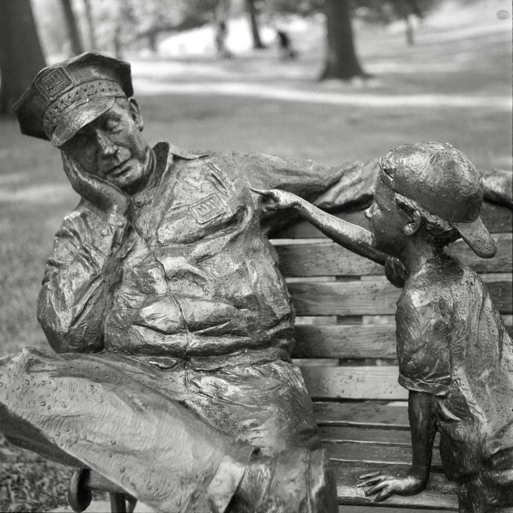 A black and white film photo. A bronze statue of a boy pointing at the resting Maytag Repairman at Maytag Park in Newton, Iowa. Saturday, July 12, 2025. 