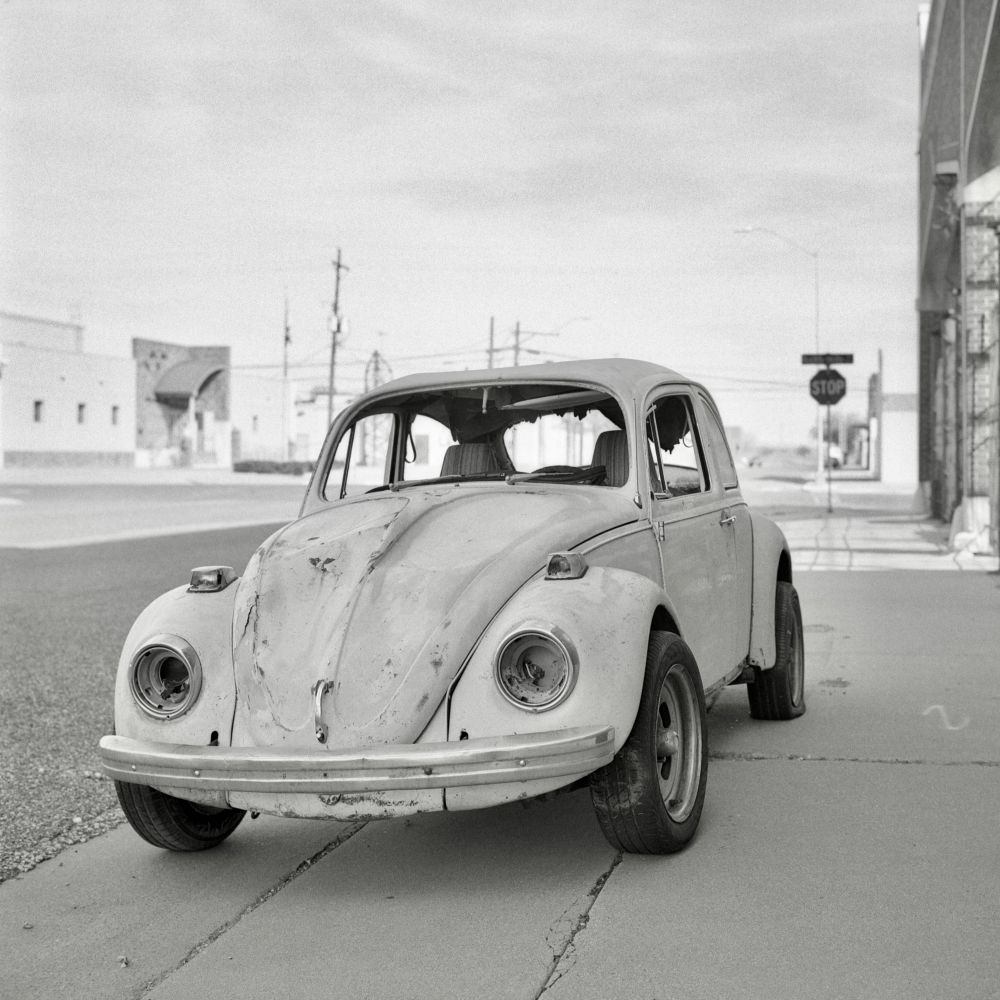 A black and white film photo. An old Volkswagen Beetle, who has a missing windshield, headlights and is dented up all over, sits quietly on a sidewalk in Lubbock, Texas. Sunday, February 16, 2025.
