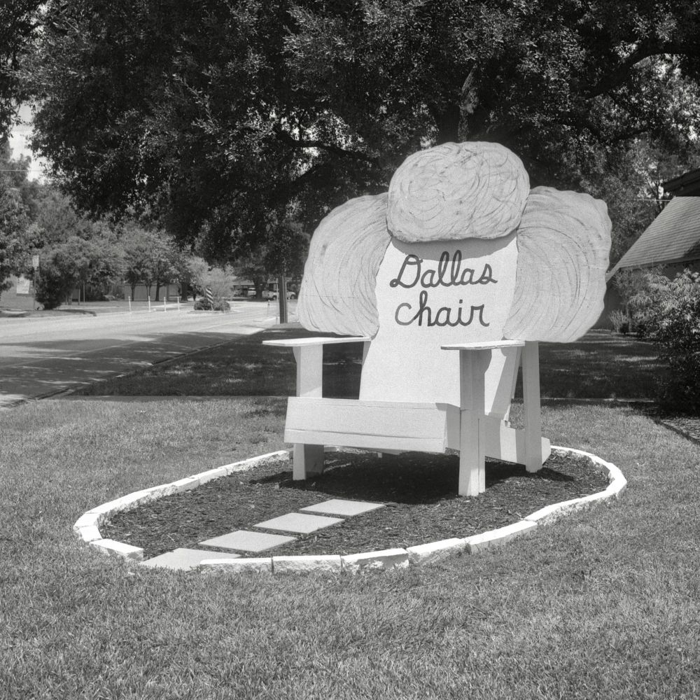 A black and white film photo. A giant wooden chair sporting an enormous hairdo with the words “Dallas Chair” is stationed outside a church in Austin, Texas. Saturday, August 2, 2025. 