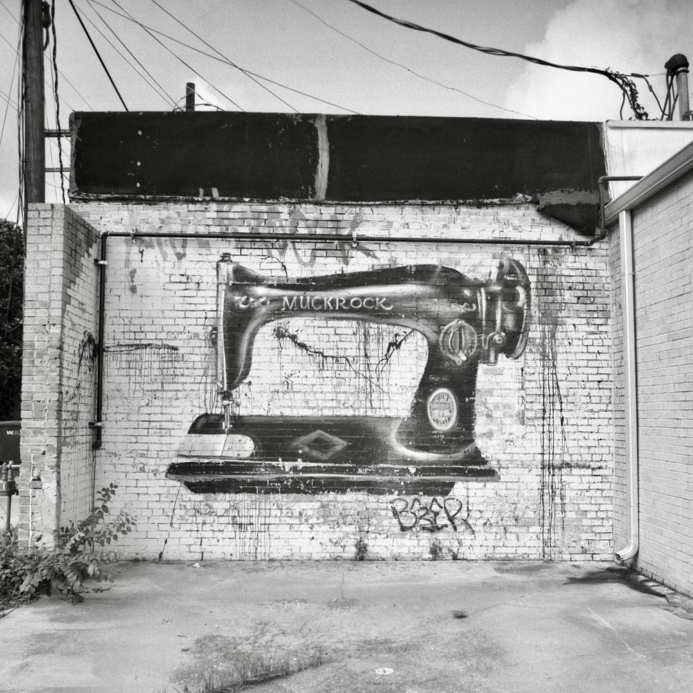A black and white film photo. Graffiti art of a giant antique sewing machine is shown on an old white brick wall in Smithville, Texas. Sunday, June 29, 2025. 