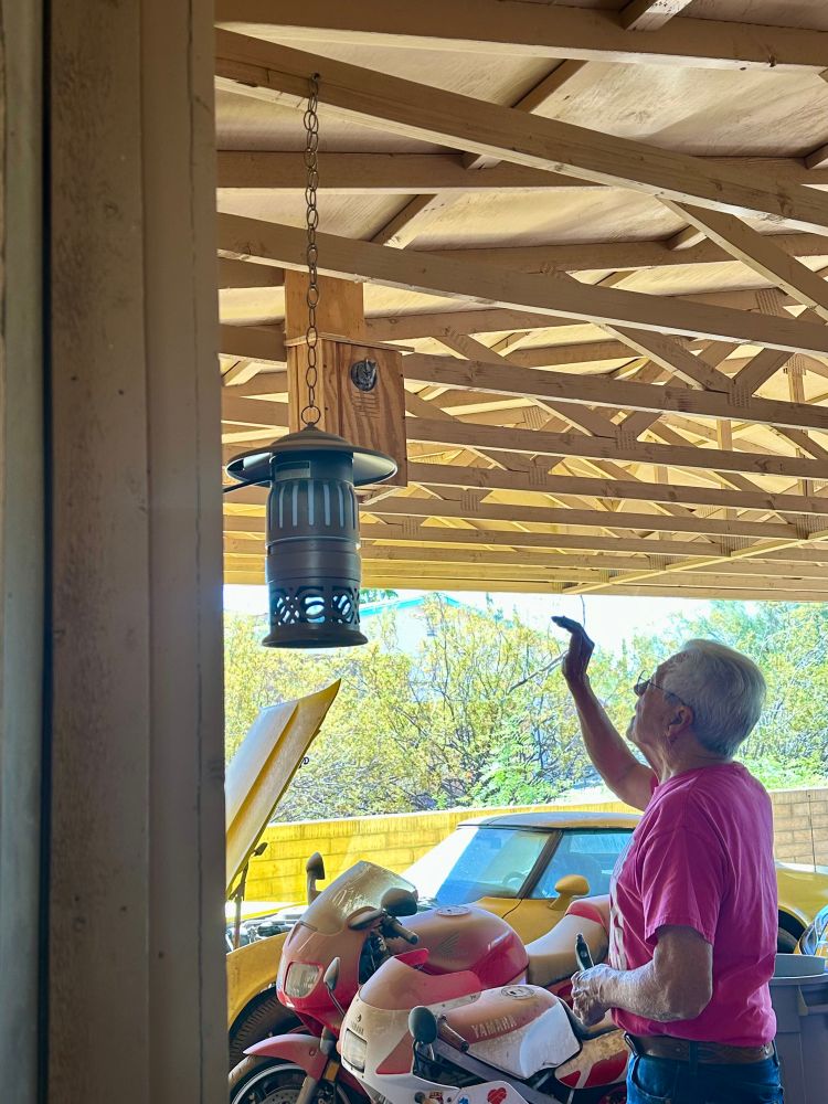 A picture of a man (my dad) waving at a western screech owl in the box he made for them. The box is in the ceiling of a carport and you can also see some of dad’s other toys in the background. Two Honda motorcycles and a 1980 Corvette(!).