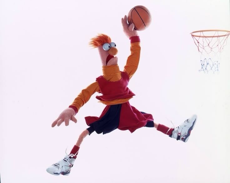 A puppet/muppet in a red basket ball jersey, black and red basket ball shorts, and white athletic shoes is dunking a basketball. He resembles the Jordan logo