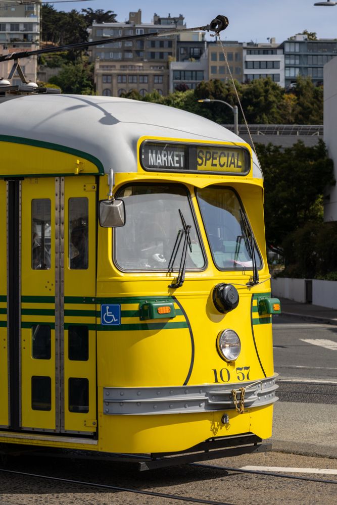 Close-up of a bright yellow vintage streetcar in San Francisco with green stripes and a sign reading “Market Special.” The front of the streetcar features rounded windows, headlight, and a mounted mirror. In the background, hillside buildings and trees are visible under a clear sky.