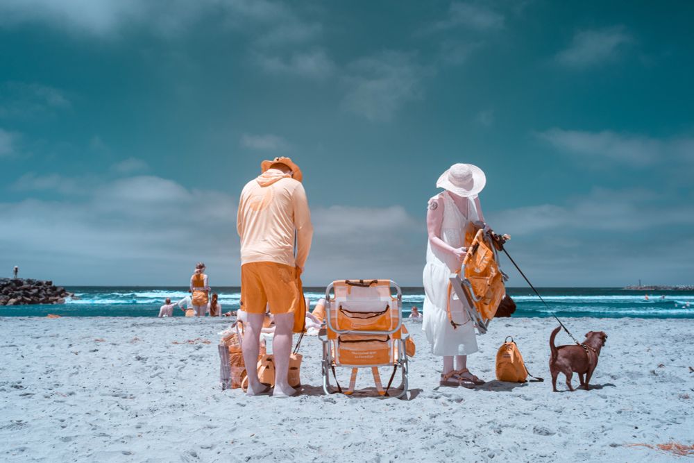 A color-swapped infrared photo of a beach scene showing a man and woman setting up orange beach chairs. The man wears orange shorts and a long-sleeve shirt with a hat, facing away. The woman, in a white dress and sunhat, holds a chair and a leash attached to a small brown dog. The sky appears teal, and the sand is pale blue. Other beachgoers and the ocean are visible in the background.