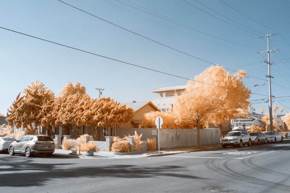 A residential street corner captured in infrared, with foliage rendered in pale orange and the sky a soft blue. Several cars are parked along the curb. A stop sign, low fencing, and a mix of single-story and multi-story buildings define the urban neighborhood scene. Power lines stretch overhead.