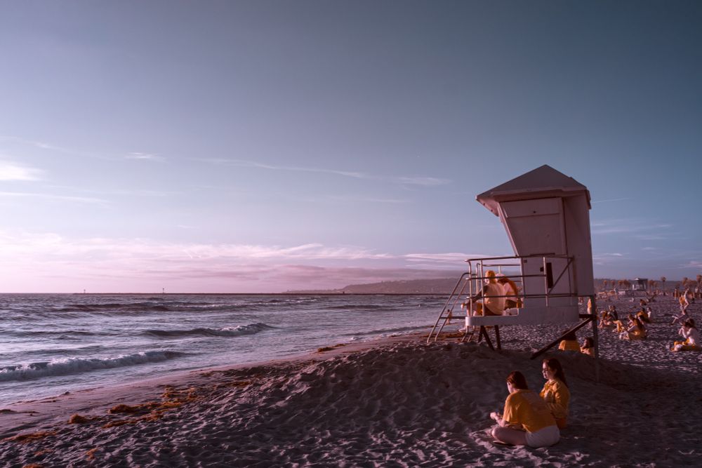 A wide infrared photograph of a Southern California beach, showing a lifeguard tower to the right and groups of people scattered along the sand. The two figures in the foreground sit cross-legged, facing each other, wearing warm-toned clothing. The ocean appears deep blue with gentle waves, and the sky fades from lavender near the horizon to teal higher up. The overall color palette is surreal but cohesive, casting the familiar beach scene in an otherworldly light.
