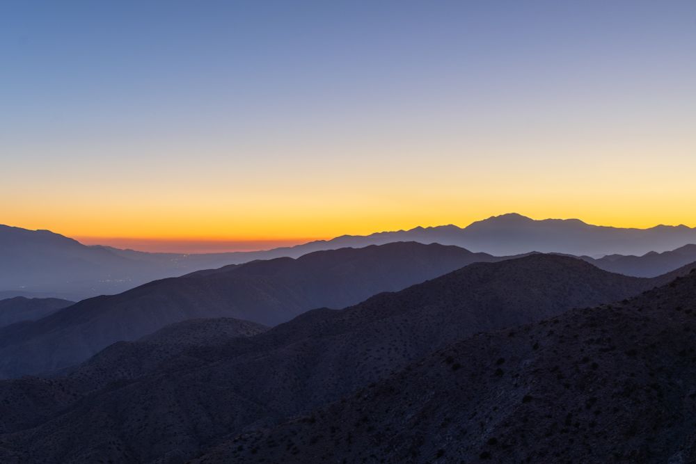 A layered mountain range at sunset with ridgelines fading into the distance. The sky shifts from a deep orange glow near the horizon to soft blue higher up. Light haze hangs in the valleys, which adds depth and atmosphere to the desert landscape.