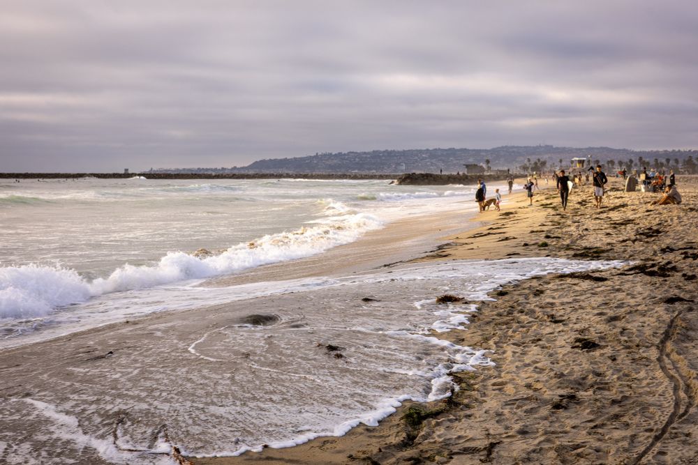 High tide peaks along the shoreline at Ocean Beach in San Diego, with waves pushing high into the sand under a soft, overcast sky. Golden evening light illuminates people walking near the water and others gathered farther up the beach. A rocky jetty stretches into the ocean, and distant hills rise on the horizon.