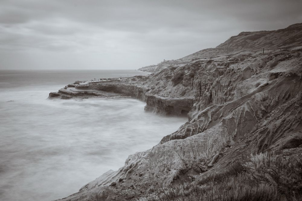 A black-and-white infrared photo of sandstone cliffs overlooking the ocean on a cloudy day. The long exposure smooths the water into a misty blur as it laps against the base of the cliffs. The layered rock formations lead the eye toward the horizon, where a few small figures stand near the edge.