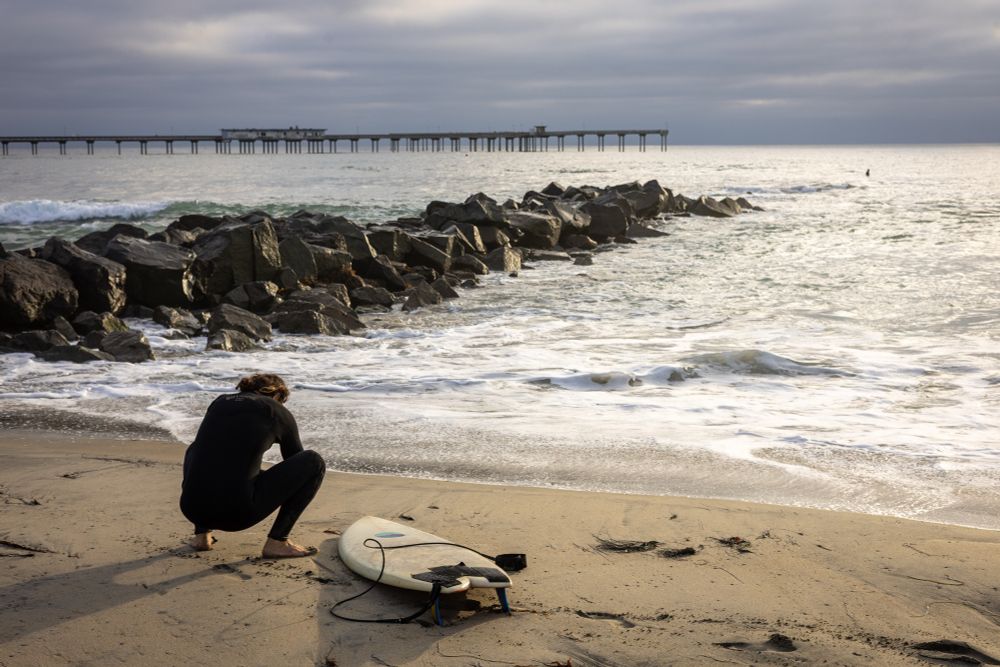 A surfer in a black wetsuit crouches barefoot on the sand near a surfboard, gazing at the ocean. Jagged rocks lead into the water, and a long pier stretches across the horizon under a cloudy sky. A lone surfer floats in the distance.